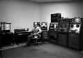 Photograph: [Larry Austin sitting in a room of equipment]