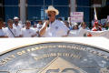 Photograph: [Speaker wearing a hat with a microphone and protesters behind him]