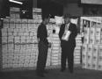 Photograph: [Two Men Standing in Front of Flour Bags]