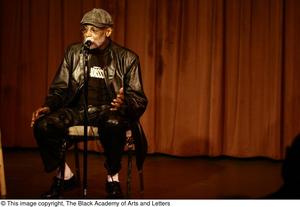 [Photograph of Melvin Van Peebles seated in a chair on stage]