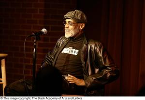 [Photograph of Melvin Van Peebles seated on a stage with a microphone]