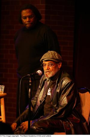 [Photograph of an unidentified man standing behind Melvin Van Peebles on stage]