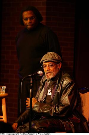 [Photograph of an unidentified man standing behind Melvin Van Peebles on a stage]