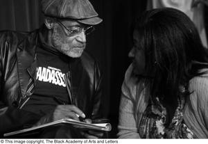 [Photograph of Melvin Van Peebles signing a book for a woman as he sits on stage]