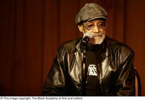 Primary view of object titled '[Photograph of Melvin Van Peebles sitting in a chair on stage]'.
