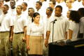 Primary view of [Choir members sing while standing around a mic]