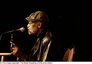 [Photograph of director Melvin Van Peebles seated on stage with a microphone]