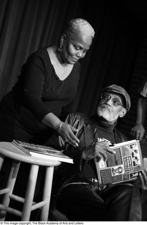 [Photograph of a woman standing next to Melvin Van Peebles as he signs books]