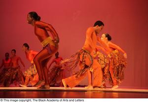 [Photograph of many men and women dancing on stage in orange outfits]