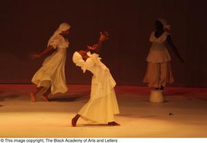 [Photograph of three women dancing on a stage in white dresses]