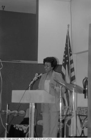 [Woman standing at podium and addressing guests at conference]