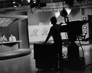 [Photograph of camera operators silhouette filming news team on set]