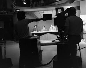 [Camera operators silhouette filming news team on set]