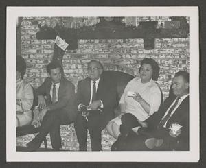 [Photograph of a group of individuals sitting in front of a fireplace]