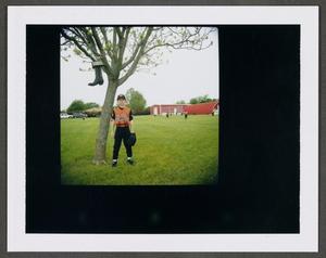 [Boy in a baseball uniform standing next to a tree]
