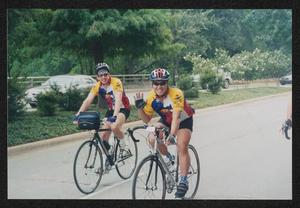 [Cyclist smiling and waving to the camera as they ride down the street: Lone Star Ride 2003 event photo]