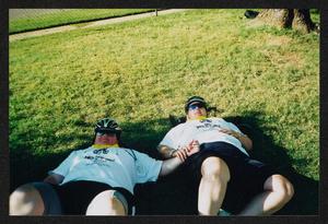 [Two cyclists laying in the grass holding arms: Lone Star Ride 2003 event photo]