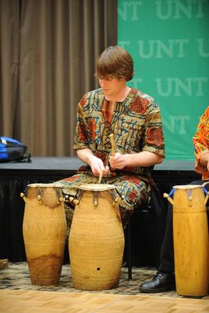 [A man in patterned clothing sitting while playing two drums]