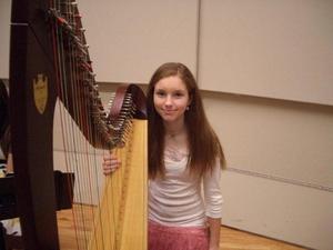 [A girl in a white shirt and pink skirt sitting behind a harp]