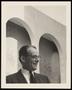 Photograph: [A man in a suit and glasses standing in front of archways]