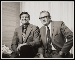 [Two men in suits smiling and posing on a desk]