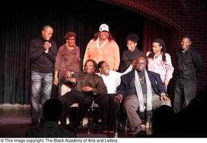 [Performer, man, and Curtis King sitting on stage with three women and three men standing]
