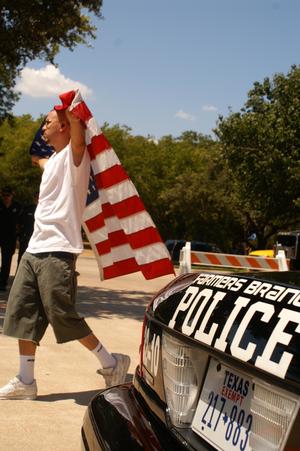 [Man with a U.S. flag walks past a police car]