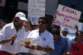 Photograph: [Two men (singing?) with a microphone and protesters behind them]